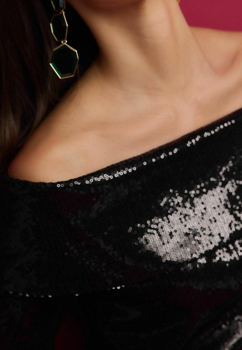 Clavicule et cou d'une personne portant un haut noir à paillettes et épaules dénudées avec une boucle d'oreille géométrique verte et dorée sur un côté.