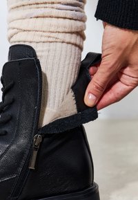 Cizmulita neagră din piele cu fermoar, având o căptușeală interioară moale și un exterior texturat. O mână ajustează deschiderea cizmei.