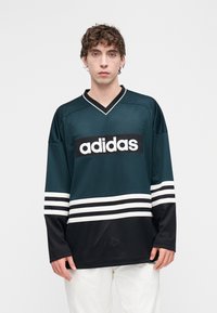 Türkizkék és fekete hálós mez, amelyen egy feltűnő fehér Adidas logó látható, V-nyakú kivágással, fekete ujjakkal és fehér csíkokkal. Texturált anyag.