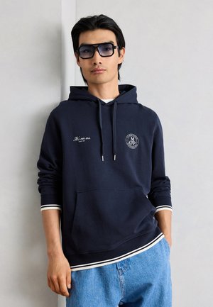 Nuori mies pukeutunut tummansiniseen huppariin, jossa valkoinen teksti ja logot, vaaleansiniset farkut ja suuret tummat aurinkolasit, seisoo vaaleaa seinää vasten.