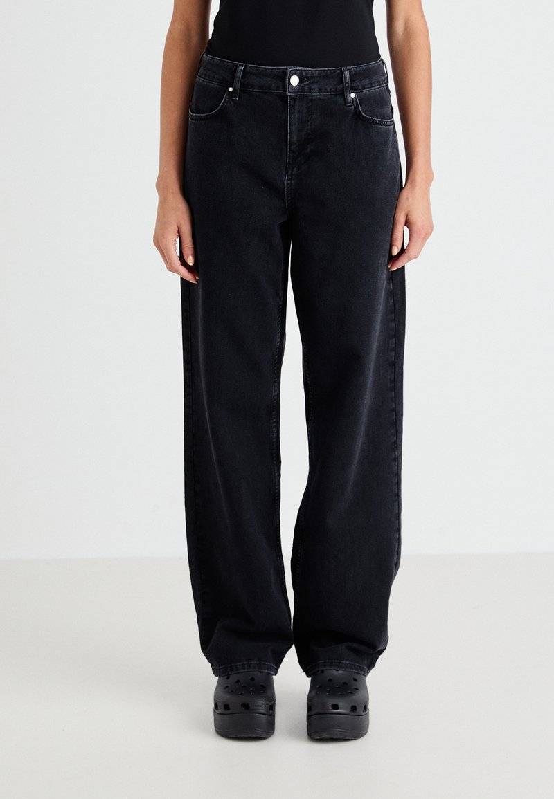 Jean en denim noir à jambes larges avec taille haute, cinq poches et texture délavée. Associé à des sabots noirs.