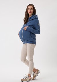 Felpa da maternità blu con zip, dotata di una grande tasca frontale e cappuccio, indossata con pantaloni fitted chiari e scarpe sportive.