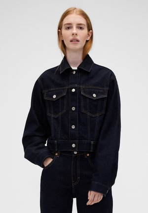 Jeune femme aux longs cheveux roux portant une veste en jean noire et un jean noir assorti sur un fond uni.