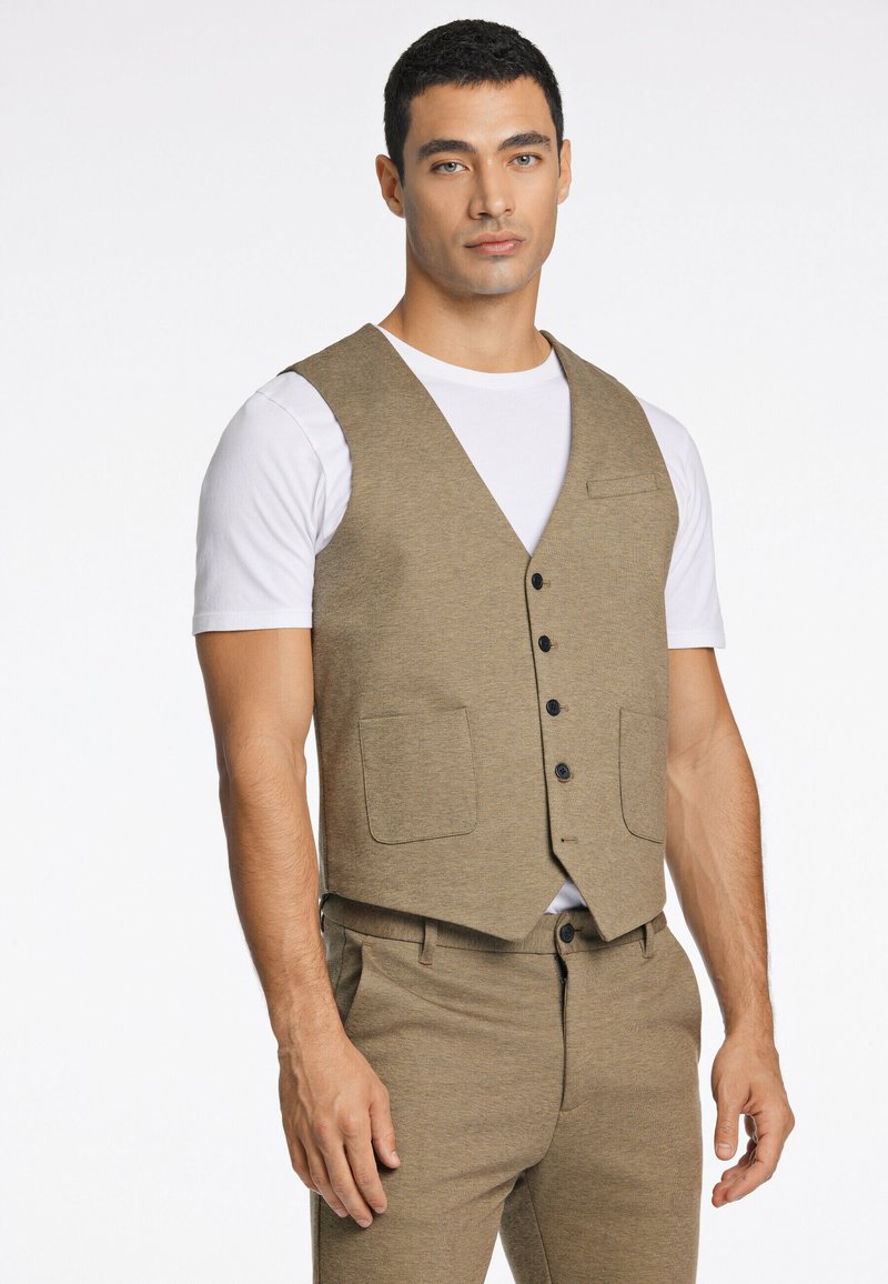 Gilet marrone con design a scollo a V, dotato di quattro bottoni neri, due tasche frontali e indossato sopra una t-shirt bianca. Il tessuto ha una finitura a trama.