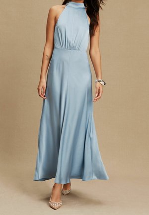 Femme portant une robe bleu clair sans manches à col halter avec une jupe fluide et des talons beiges ornés de perles.