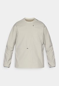 Veste beige clair en matériau résistant à l'eau. Elle possède des manches longues, un col rond et une poche frontale avec une fermeture éclair.