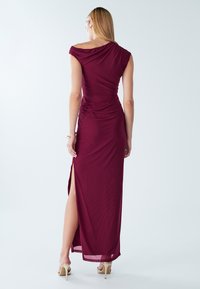 Vīna sarkanas krāsas gara kleita ar drapētu kakla izgriezumu un savāktu detaļu pie vidukļa, ar sānu šķēlumu un gludu, elegantu audumu.