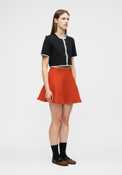 Νεαρή γυναίκα που φοράει μαύρο κοντομάνικο κοντό ζακετάκι με λευκή επένδυση, πορτοκαλί πιετέ φούστα, μαύρες κάλτσες και μπαλαρίνες με σχέδιο λεοπάρ.