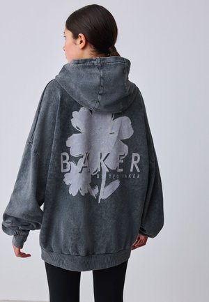 Žena s tmavými vlasmi v nízkom chvoste, oblečená v oversize vyblednutej čiernej mikine s veľkým bielym kvetinovým vzorom a nápisom „BAKER TED BAKER“ na chrbte.