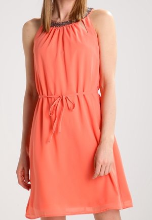 Vrouw draagt een mouwloze koraalkleurige jurk met een met kralen versierde halslijn en een dunne tailleband, staand tegen een effen lichte achtergrond.