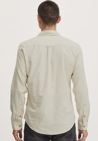 Chemise à manches longues beige clair en tissu texturé avec un devant à boutons, dotée d'un empiècement au dos et de poignets à boutons.