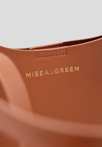 Sac à main en cuir marron avec une texture lisse, arborant un lettrage doré en relief "MISEAUGREEN" et un design minimaliste.