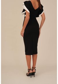 Svart klänning med vita volanger i korta ärmar, figurnära silhuett, knä-lång, slits bak och V-formad ryggdesign. Kombineras med svarta klackar.