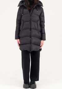 Zwarte pufferjas met een capuchon, voorzien van een ritssluiting aan de voorkant, doorstikte textuur en verstelbare riemen. Draagt met zwarte broek en chunky schoenen.