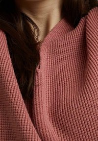 Pull rose en maille gaufrée avec un décolleté en V profond et un léger recouvrement au niveau du col, mettant en valeur un tissu tricoté doux.