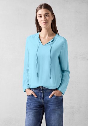 Haut long à manches bleu clair avec un col en V et une capuche à cordon. Tissu texturé associé à un jean en denim, les mains dans les poches.