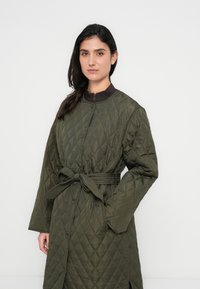 Πρασινωπό μπουφάν με καπιτονέ σχέδιο, κλείσιμο με κουμπιά, ζώνη στη μέση και πλαϊνές τσέπες. Διαθέτει ψηλό γιακά και μοτίβο διαμαντιών σε όλο το μήκος.