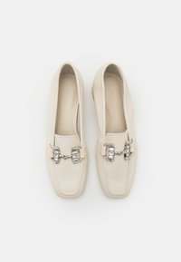 Mocassins en cuir couleur crème avec des bouts carrés et des chaînes décoratives argentées sur le devant, vus de dessus sur un fond blanc.