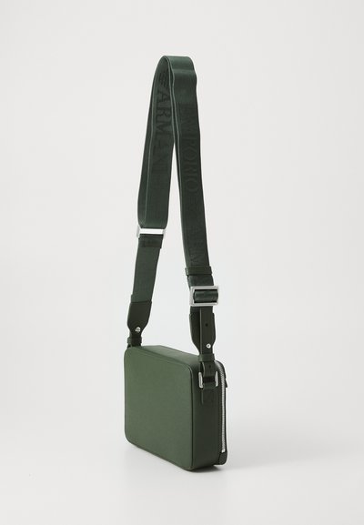Grønn stoff crossbody-veske med rektangulær form, teksturert overflate, justerbar stropp og glidelås som har merkede detaljer.