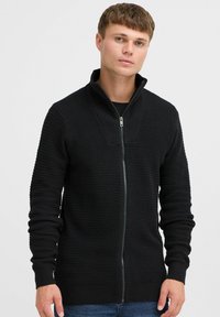 Schwarzer, melierter Strickpullover mit Reißverschluss und hohem Kragen, geripptem Muster und enganliegenden Ärmeln. Der Stoff hat ein dezentes Waffelmuster.