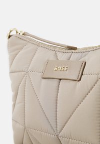 Bolso acolchado beige con detalles en dorado, que presenta una etiqueta rectangular grabada con "BOSS", resaltando su textura suave y su patrón geométrico.