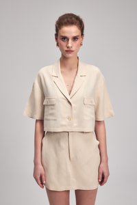 Beige zkrácená bunda s krátkými rukávy, dvěma našitými kapsami a zapínáním na knoflíky, doplněná odpovídajícími sukněmi s vysokým pasem. Hladká textilie.