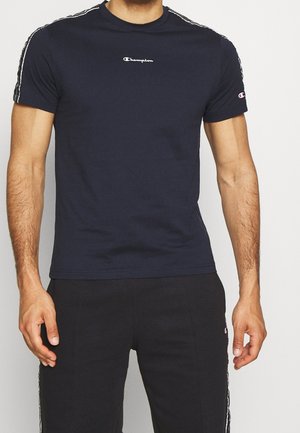 Muž v tmavě modrém tričku Champion s logem na hrudi a pruhovaných černých kalhotách Champion, stojící se uvolněně spuštěnými pažemi podél těla.