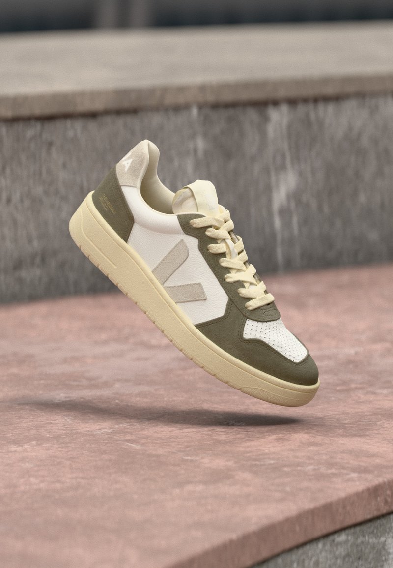 Χαμηλό παπούτσι τύπου sneaker με λευκή διάτρητη μύτη, λεπτομέρειες από σουέντ ελιάς, κρεμ κορδόνια και μπεζ σόλα που αιωρείται πάνω από ροζ πέτρινη επιφάνεια.