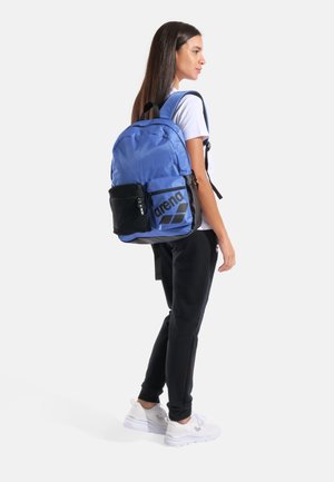 Sac à dos bleu avec un logo noir, doté d'une poche en filet sur le côté, de bretelles ajustables et d'une texture de tissu lisse et résistante.