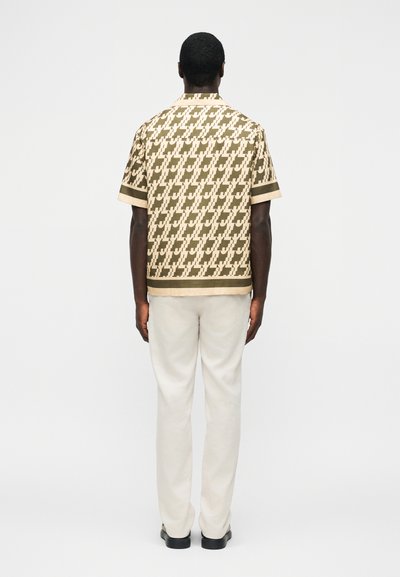 Άνδρας που φοράει μπεζ κοντομάνικο πουκάμισο με γεωμετρικό σχέδιο, κρεμ παντελόνι και μαύρα παπούτσια, στέκεται με την πλάτη προς τα εμπρός σε λευκό φόντο.