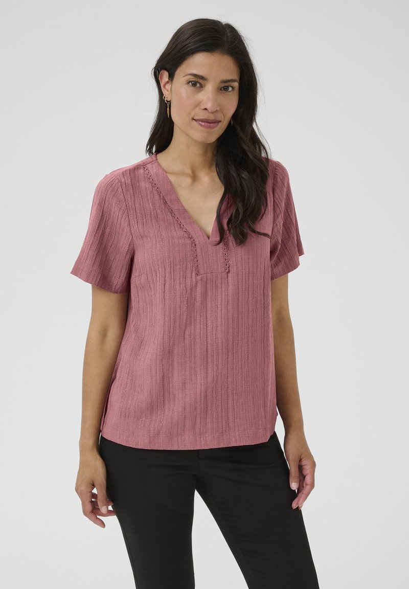 Blouse à manches courtes et col en V dans une couleur rose texturée. Présente des détails en dentelle le long de l'encolure, avec des fentes sur les côtés pour plus de mouvement.