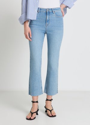 Donna che indossa jeans svasati corti azzurri con orli sfrangiati, sandali neri con tacco e cinturini alla caviglia e una camicia a righe blu e bianca.