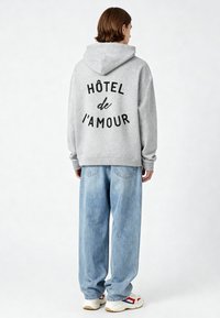 Szürke kapucnis pulóver, amelyen hátul nagy fekete betűkkel a "HÔTEL de l'amour" felirat található, világoskék bő nadrággal és fehér sneaker cipővel párosítva.