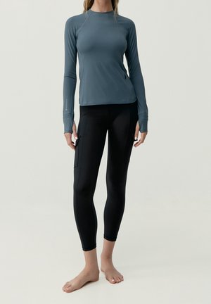 Kobieta ubrana w dopasowany, długi rękaw w kolorze zielonkawoniebieskim oraz czarne krótkie legginsy z bocznymi kieszeniami, stojąca boso na jasnym tle.