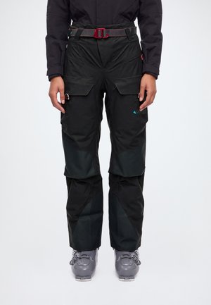 Modèle portant un pantalon de ski technique noir, une ceinture à boucle rouge et des chaussures de ski grises sur un fond blanc de studio.