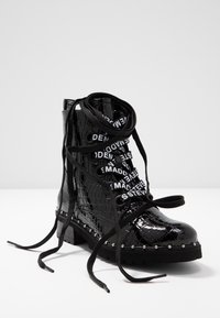 Bottine en cuir verni noir à texture crocodile, clous décoratifs argentés le long de la semelle, et lacets de marque noirs et blancs noués lâchement.