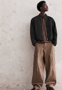 Crna bomber jakna sa patentnim zatvaračem, smeđa karirana košulja i široke smeđe cargo hlače s velikim džepovima. Glatke teksture tkanina i neutralne boje.