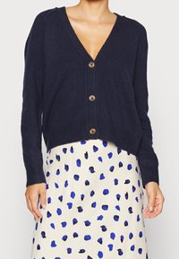Navy cardigan with a V-neck, long sleeves, and wooden buttons. Worn over a cream skirt with blue polka dots, showcasing a relaxed fit.