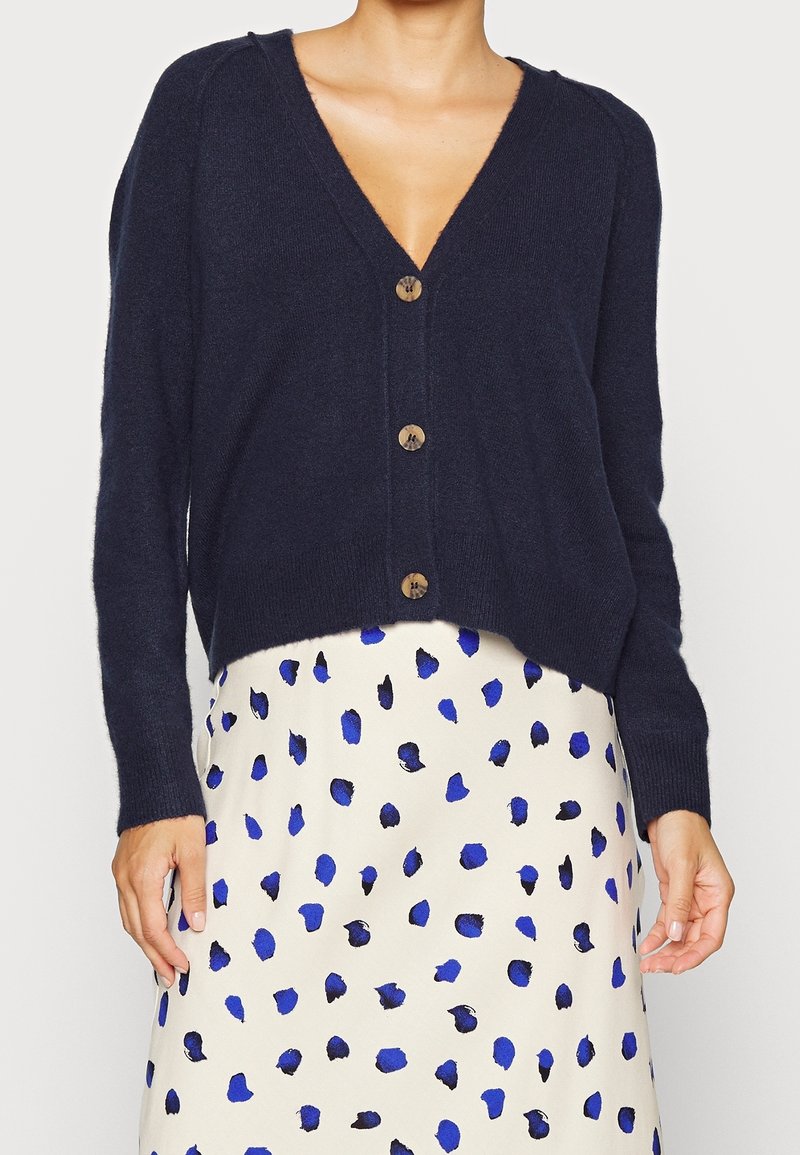 Navy cardigan with a V-neck, long sleeves, and wooden buttons. Worn over a cream skirt with blue polka dots, showcasing a relaxed fit.