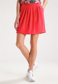 Plisovaná sukně ve tvaru A v jasně korálové barvě, s texturovaným povrchem. S kombinovanými bílými teniskami.