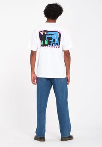 White t-shirt featuring a colorful graphic on the back, depicting abstract shapes in blue, green, and pink, paired with loose denim jeans.