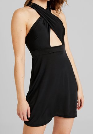 Femme portant une robe noire sans manches avec un col halter torsadé et un détail découpé à l'avant sur un fond uni clair.