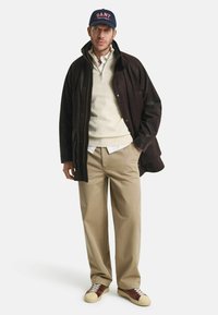 Leren bruine suede jas, crèmekleurige gebreide trui, witte blouse, khaki broek en bruine leren sneakers met lichtgekleurde zolen, marineblauwe pet met logo.
