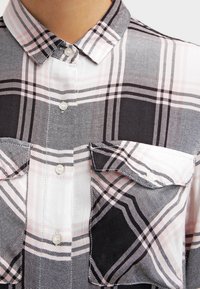 Camicia a quadri con colletto bottonato, tessuto grigio e bianco, dettagli rosa e due tasche sul petto. Materiale testurizzato con un motivo a scacchi.