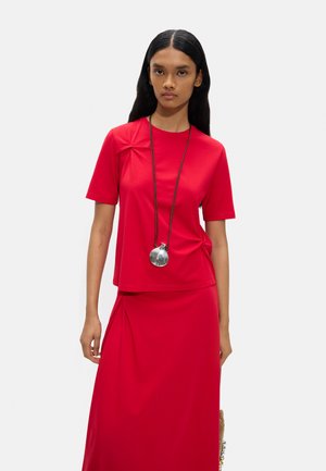 Kobieta z długimi czarnymi włosami nosi czerwoną bluzkę z krótkim rękawem i pasującą spódnicę, uzupełnione długim naszyjnikiem z dużym srebrnym wisiorkiem.