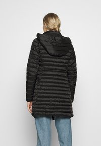 Schwarze Pufferjacke mit horizontal gestepptem Muster, Kapuze und leicht taillierter Passform. Kombiniert mit hellblauen Jeans.