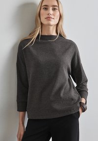 Dunkelgrauer, strukturierter Pullover mit hohem Kragen und Dreiviertelärmeln. Getragen mit schwarzen Hosen, die eine lockere Passform und minimalistische Details aufweisen.