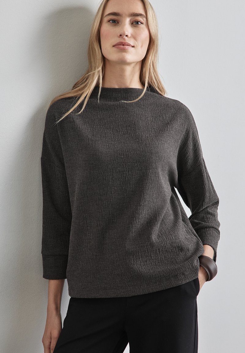 Dunkelgrauer, strukturierter Pullover mit hohem Kragen und Dreiviertelärmeln. Getragen mit schwarzen Hosen, die eine lockere Passform und minimalistische Details aufweisen.