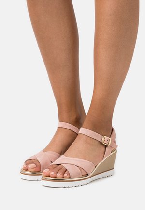 Des pieds portant des sandales compensées en suède rose clair avec des sangles à la cheville et une boucle en or, une peau lisse marron clair, des ongles de pieds bien entretenus.