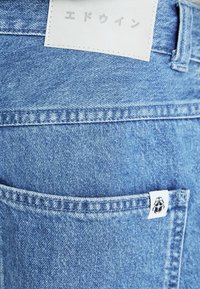 Jean en denim bleu clair avec une poche arrière visible et une étiquette blanche sewn à la taille. Le tissu a une texture délavée et un détail de couture.